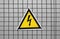 High voltage triangle warning sign mounted on a metal lattice fence on a gray background.