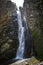 A high waterfall with white splashes of water descends from the mountain and sparkles in the sun.