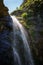 A high waterfall with white splashes of water descends from the mountain and sparkles in the sun.