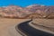 A highway curving through a barren desert landscape towards a range of colorful rugged mountains