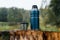 Hike, camping concept. Thermos and aluminum mug hot drink with rising steam standing on stump in rainy weather in nature