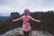 Hiker female in outdoor clothing raised hands on the top of a mountain and enjoying view of cliffs ridge and wild forest