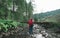 Hiker girl stands on stones of mountain stream on background of fallen tree trunk and looks at scenery. Woman in red raincoat on a