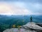 Hiker in green windcheater, cap and dark trekking trousers stand on mountain peak rock.