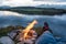 Hiker with hiking boots are relaxing by bonfire outdoors with lake and mountain view