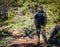 Hiker man photographer in camouflage outfit with a backpack and tripod standing on a mountain forest trail and watching wildlife,