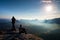 Hiker and photo enthusiast stay with tripod on cliff and thinking. Dreamy fogy landscape, blue misty sunrise in a beautiful valley