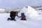 Hiker pours tea from his thermos to his friend sitting in a snowy house igloo