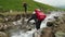 A hiker with a red cape on her backpack crossing a mountain stream, helping herself with trekking poles. In the