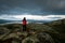 A hiker with red jacket and his dog are standing on a mountain top looking towards lakes and dramatic stormy clouds during blue ho