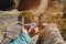 Hiker sitting on a high rock holds a compass in front of her feet in trekking boots and a high cliff with an asphalt