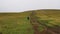 Hikers with backpacks climb a hill covered with grass. People walk along the dirt road. In the background is a gray cloudy sky. Th