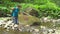 Hiking beautiful woman with backpack balancing hands crosses a mountain river with large boulders and green moss in