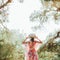 A hippie girl in a beautiful flying dress, hat and round glasses in a pine forest among the mountains.