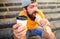 Hipster bite hot dog hold drink paper cup. Man bearded shows paper cup drink stairs background. Energy from street food