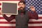 Hipster chic. Bearded hipster holding scissors and blackboard in class. Hipster man on american flag background. Hipster