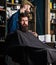 Hipster client getting haircut. Haircut process concept. Man with beard covered with black cape waiting while barber