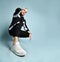 Hipster fellow in cap, black tracksuit, bracelet and sneakers. He is squatting and smiling, posing on blue background. Close up