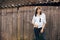 Hipster girl in white casual outfit posing on background of old wooden cabin in mountains. Stylish woman in hat standing near old