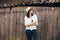 Hipster girl in white casual outfit posing on background of old wooden cabin in mountains. Stylish woman in hat standing near old