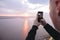 Hipster traveler taking photo on phone of amazing sunset view on river while standing on top of rock mountain. Atmospheric  moment