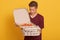 Hirizontal shot of handsome man wearing burgundy casual t shirt, holding stack of pizza boxes in hands, looking at opened box,