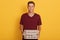 Hirizontal shot of handsome man wearing maroon casual t shirt, holding pizza boxes in hands, looking directly at camera, posing