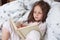 Hirizontal shot of happy girl lying in bed on blanket, reading interesting book,has concentrated facial expression, female child