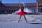 Hispanic adult female classical ballet dancer in red tutu doing figures in the middle of a plaza on a sunny day with a water