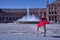 Hispanic adult female classical ballet dancer in red tutu doing figures in the middle of a plaza on a sunny day with a water