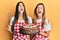 Hispanic family of mother and daughter wearing baker apron holding homemade cake angry and mad screaming frustrated and furious,