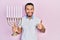 Hispanic man with beard holding menorah hanukkah jewish candle smiling happy and positive, thumb up doing excellent and approval