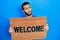 Hispanic man with beard holding welcome doormat in shock face, looking skeptical and sarcastic, surprised with open mouth