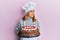 Hispanic young chef woman holding chocolate cake smiling looking to the side and staring away thinking