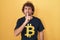 Hispanic young man wearing bitcoin t shirt asking to be quiet with finger on lips