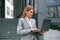 Holding laptop in hands, standing. Woman in formal clothes is outdoors near the business building