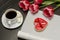 Holiday concept. Bouquet of pink tulips, a cup of coffee, red heart-shaped cookies with a note, empty magazine on a black wooden