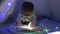 Home education, boy with glasses is holding a mobile phone with flashlight in his hands and is reading book in tent