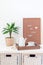 Home interior with white chest of drawers, green palm in  pot, wooden tray with tea, and pegboard with the inspiring phrase,