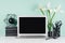 Home workspace with blank laptop monitor, black stationery, books, modern sculpture, coffee cup, white flowers in green mint.