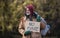 Homeless beggar man standing outdoors in park, holding bag and cardboard sign.