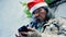 A homeless elderly Asian man wearing red christmas hat stands next to wall using his mobile phone to seek work and government