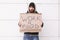 A homeless man with a beard and in a hood stands with a sign. Outdoor.