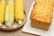 Homemade Brazilian corn bread on a cutting board and raw corn cobs in a woven bamboo basket. Isolated on jute background. Close-up