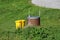 Homemade brown colored well with locked shiny metal cover next to two bright yellow plastic garbage cans surrounded with uncut