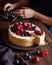 Homemade creamy mascarpone New York Cheesecake with berries on dark wooden table. Hands in the frame.