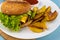 Homemade imperfect burger with potatoes and salad on a plate. Food on the table in a white plate on a wooden board.