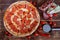 Homemade spicy pizza with pepperoni and salami lies on a wooden surface next to a special circular knife, tomatoes, red pepper,