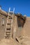 Homemade wooden ladder against side of mud adobe pueblo house where tar paper is being put on roof - with dramatic shadows under