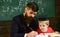 Homeschooling concept. Teacher in formal wear and pupil in mortarboard in classroom, chalkboard on background. Father